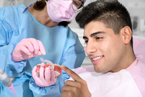 A dental professional wearing a blue gown, pink gloves, and a pink mask holds a dental model alongside an orthodontic appliance, likely a clear aligner or retainer. A patient points at the model while the professional explains or demonstrates the use of the appliance in a clinical setting.